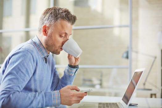 Businessman in casualwear having tea while reading sms on cellphone in office
