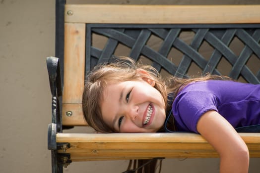 Little girl lying on her stomach on an outside bench with a big smile on her face.
