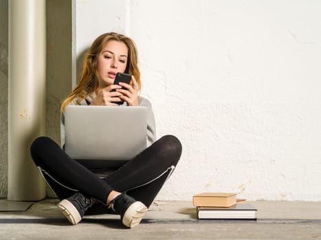 Photo of a young woman student sitting in a hallway with a laptop and looking at her smartphone.