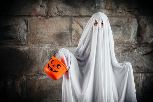 Young boy dressed in a bed sheet to be a ghost, and holding a pumpkin pail for halloween.