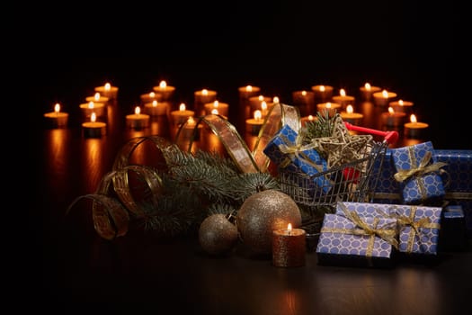 Shopping trolley, gift boxes and Christmas decoration with burning candles and fir tree branch on black background with copy space, Christmas and New Year shopping. Winter sale.