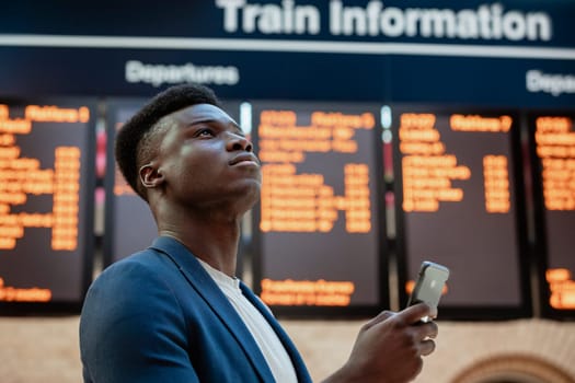 A businessman looking up in a train station while holding his mobile phone. The arrival departure board is behind him.