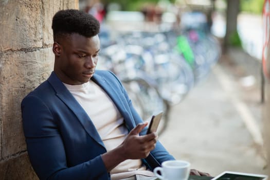Businessman sitting down at an outdoor table in a city while typing on his mobile phone.
