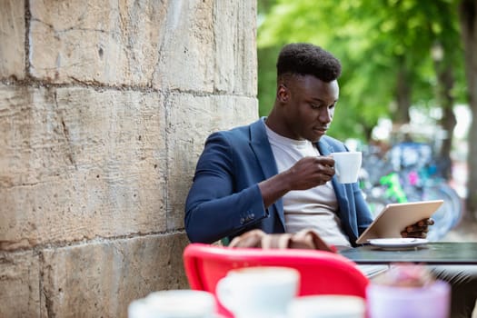 Businessman sitting down at an outdoor table in a city while drinking a hot drink and looking at his digital tablet.