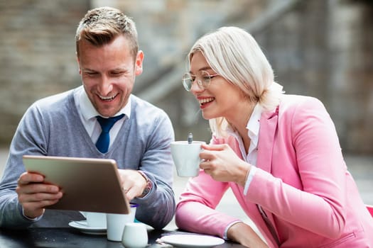 Two businesspeople sitting down and laughing at something on a digital tablet. The businesswoman is holding a hot drink while the businessman holds the tablet.