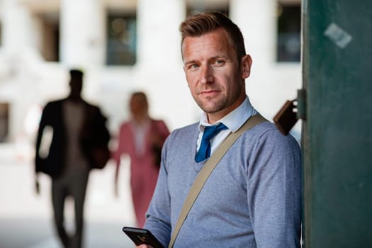 A businessman leaning against a wall while looking away from the camera with a mobile phone in his hand.