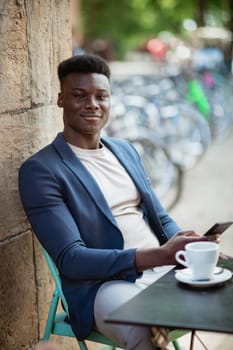Businessman sitting down at an outdoor table in a city while typing on his mobile phone and looking at the camera.