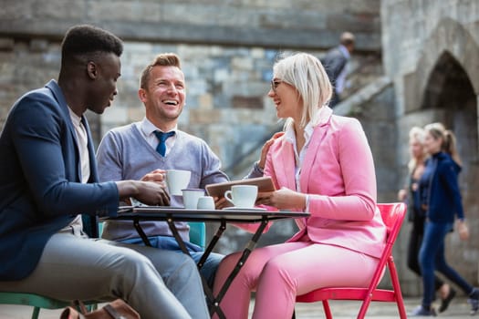 Two businessmen and a businesswoman sitting at an outdoor table in a city having a hot drink while laughing at something on a digital tablet. One man is touching the woman on the shoulder.