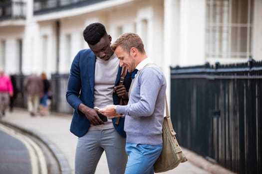 Two businessmen standing on the pavement of a city street while looking at one of their phones.
