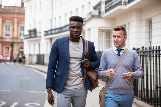 Two businessmen walking, on their way to work. They are talking and one man is holding a mobile phone ready to use it.