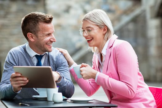 Two businesspeople sitting down and laughing at something on a digital tablet. The businesswoman is holding a hot drink and has her hand on the businessman's shoulder while he holds the tablet.