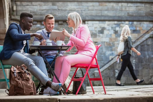 Two businessmen and a businesswoman sitting at an outdoor table in a city having a hot drink while discussing work on a digital tablet.