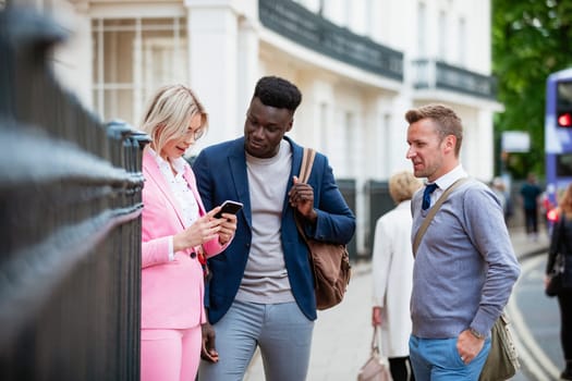 A businesswoman and two businessmen standing together on a city street. The woman is showing one man something on her phone while the other man is looking over them and smiling.