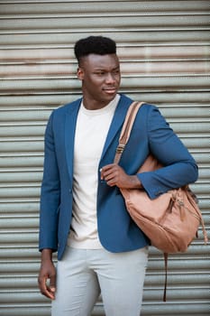Three quarter length shot of a businessman standing in front of a shutter in the street. He is wearing smart casual clothing while holding a satchel over his shoulder. He is looking to his side.