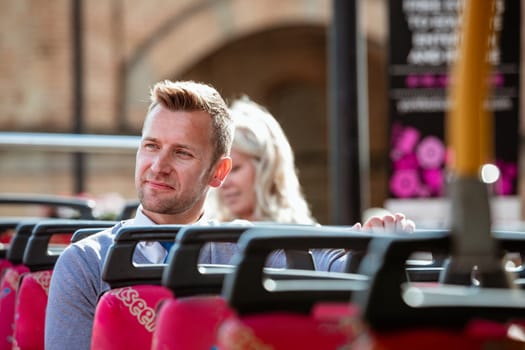 A businessman sitting by himself on an open-air bus  admiring the view. There is an unrecognisable woman behind him.
