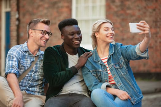 A woman taking a selfie with two other men while sitting down on a bench.