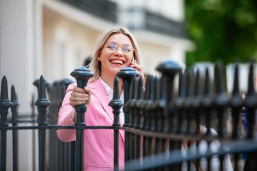 A businesswoman laughing while talking on her mobile phone. She is holding onto iron fencing.