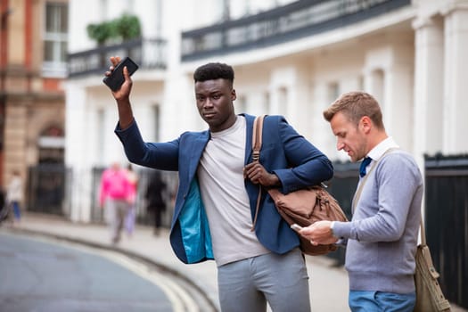 Two men trying to catch a taxi to get to work. One is trying to track one down while the other man browses on his phone.