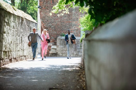 A front view shot of three businesspeople in a city street. Two are holding hands and running towards the camera while another man walks slightly behind them with a mobile phone in one hand. They are all wearing businesswear.