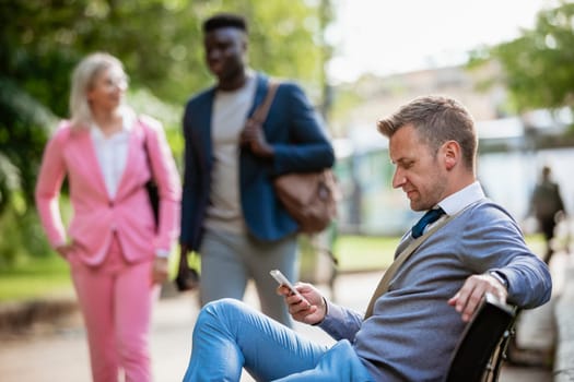 A businessman sitting on a park bench while holding and typing on his mobile phone. There are other businesspeople in the background, about to walk past.
