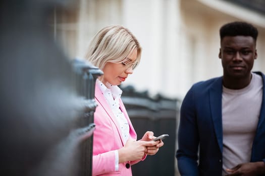 A close-up of a woman standing against an iron gate while texting on her mobile phone. A man is about to walk past her.