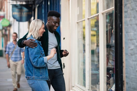 A man and woman standing outside a shop window together, he has his arm around her.