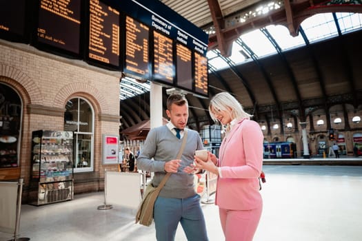 A businessman and businesswoman standing together at a train station. The woman is showing the man something on her mobile phone.