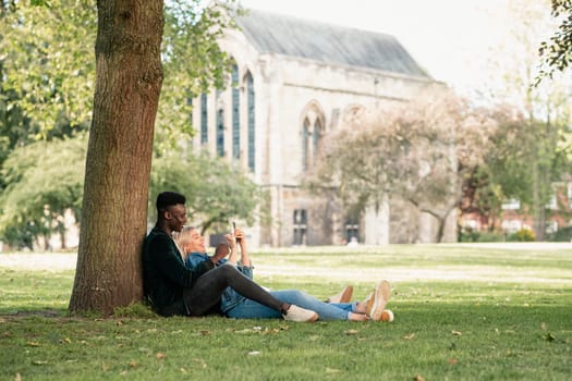 A man and woman sitting in a park against a tree. The woman is sitting in between the man's legs while showing him something on her mobile phone.