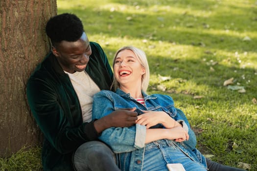 A man and woman sitting in a park against a tree. The man is holding her as she sits in between his legs. They are looking at each other while laughing.