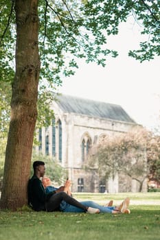 A man and woman sitting in a park against a tree. The woman is sitting in between the man's legs while showing him something on her mobile phone. The woman is looking up at the man and laughing.
