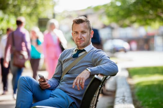 A businessman sitting on a park bench while holding his mobile phone but looking at the camera.