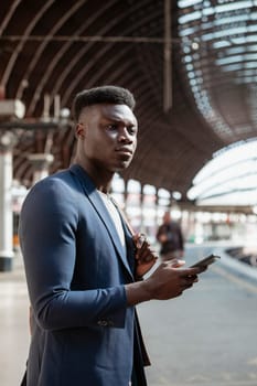 A businessman waiting for a train on the platform in a train station. He is holding a backpack over his shoulder while also holding a mobile phone in another hand.