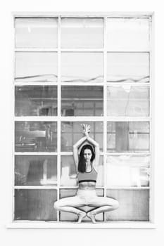 Fit sporty active girl in fashion sportswear doing yoga fitness exercise in front of big industrial window frame. colorful reflections in window glass. Urban style yoga. Black and white.