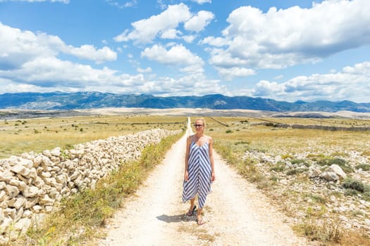 Caucasian young woman in summer dress holding bouquet of lavender flowers while walking outdoor through dry rocky Mediterranean Croatian coast lanscape on Pag island in summertime.