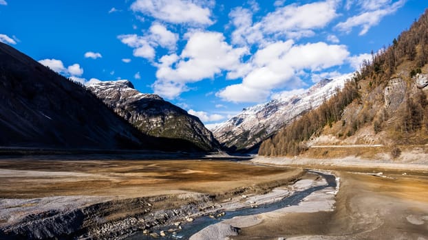 Beautiful mountain valley with stream and riverbed, trees and dried Livigno lake, puffy clouds and blue sky in background, Alps, Italy