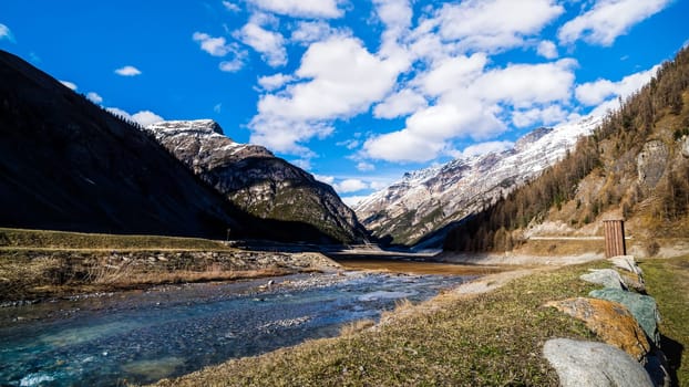 Beautiful mountain valley with stream, trees, small hut and dried Livigno lake, puffy clouds and blue sky in background, Alps, Italy