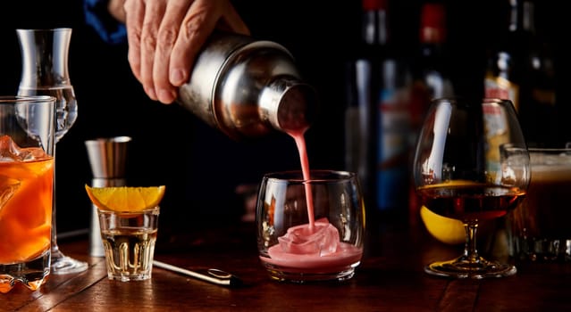 Bartender pouring a fruity cocktail from a shaker on a wooden bar counter with an assortment of different alcoholic beverages in glasses