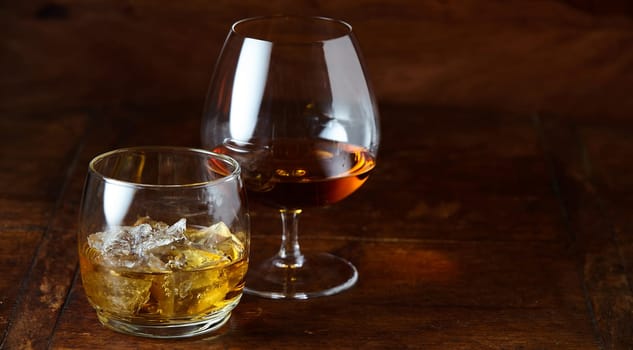 Whisky on the rocks and cognac in a snifter standing on a wooden bar counter at night with copy space