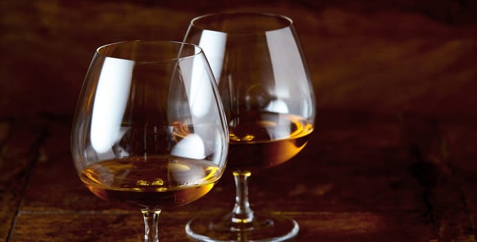 Two brandy snifter glasses with cognac viewed in close-up against dark brown background of wooden counter