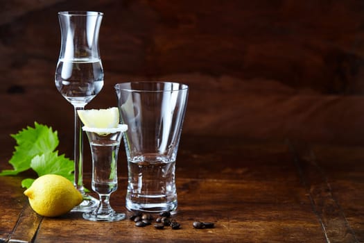 Different glasses and shots of grappa beverage on dark brown lacquered bar counter, served with lemon and decorated with green grape leaf and coffee beans. Side view with copy space
