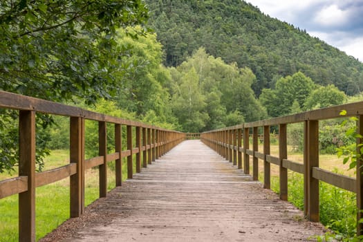Bridge made of wooden boards over a green field with trees and wooded hills