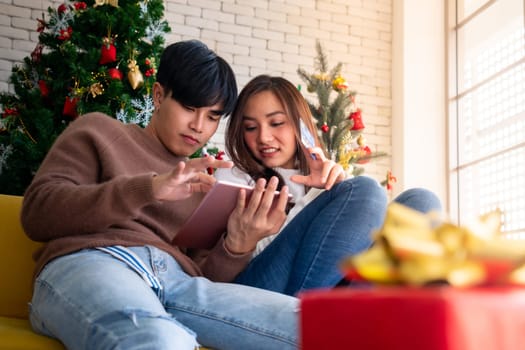 Young Adult teenager couple using tablet on sofa in living room for online shopping with gift box with Christmas tree in background