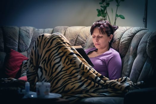 middle-aged woman is reading a book at home with the lamp on. Authentic photo, real people in real situation