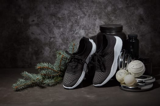 Sports nutrition (supplements), dumbbell, sneakers, branch of fir and Christmas ornaments on dark grey background with copy space. Fitness, sport or healthy lifestyle concept. New Year and Christmas.