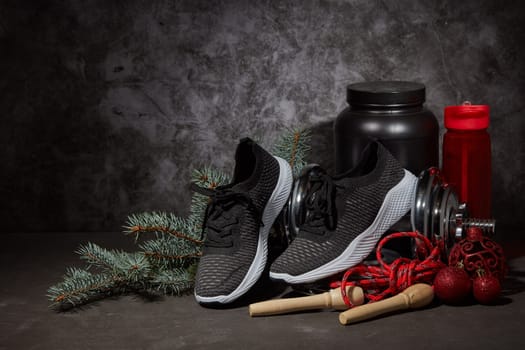 Sports nutrition (supplements), dumbbell, sneakers, jump rope and Christmas ornaments on dark grey background with copy space. Fitness, sport or healthy lifestyle concept. New Year and Christmas.