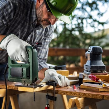 Adult carpenter craftsman wearing helmet and protective leather gloves, with electric saw working on cutting a wooden table. Construction industry, housework do it yourself. Stock photography.