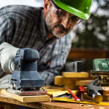 Adult carpenter craftsman wears protective leather gloves, with the electric sander smoothes a wooden table. Construction industry, housework do it yourself. Safety at work.