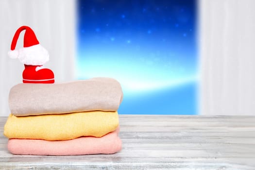 Stack of winter clothes. Pile of colorful knitted cozy warm cashmere sweaters with Santa shoe and hat above on wooden table against abstract winter background. Template for winter christmas sale.