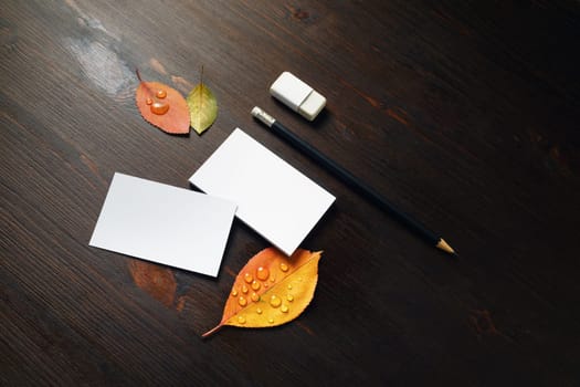 Blank business cards, pencil, eraser and bright autumn leaves on wooden background.
