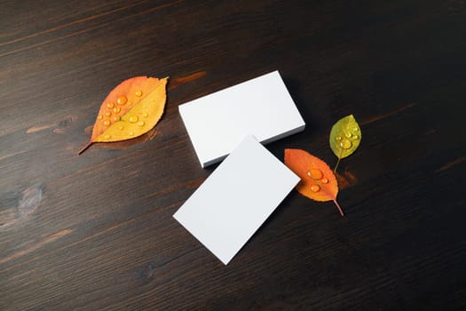Photo of blank white business cards and autumn leaves on wooden background. Copy space for text.